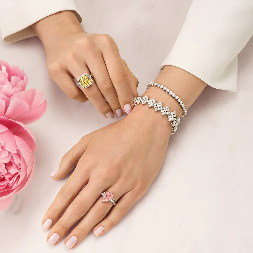 A woman's hand featured with diamond engagement and bridal jewelry by Harry Winston with a pink flower. 